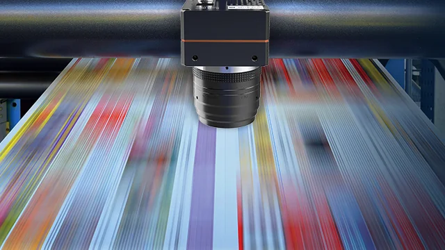Hikrobot line scan camera mounted above a fast-moving printed paper web in a printing press, capturing the material line by line